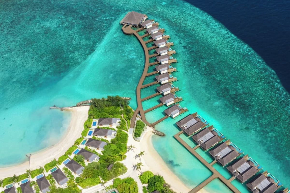 Vue aérienne d’un complexe hôtelier doté de bungalows sur pilotis reliés par des passerelles, d’une plage avec des piscines et d’une eau turquoise cristalline.