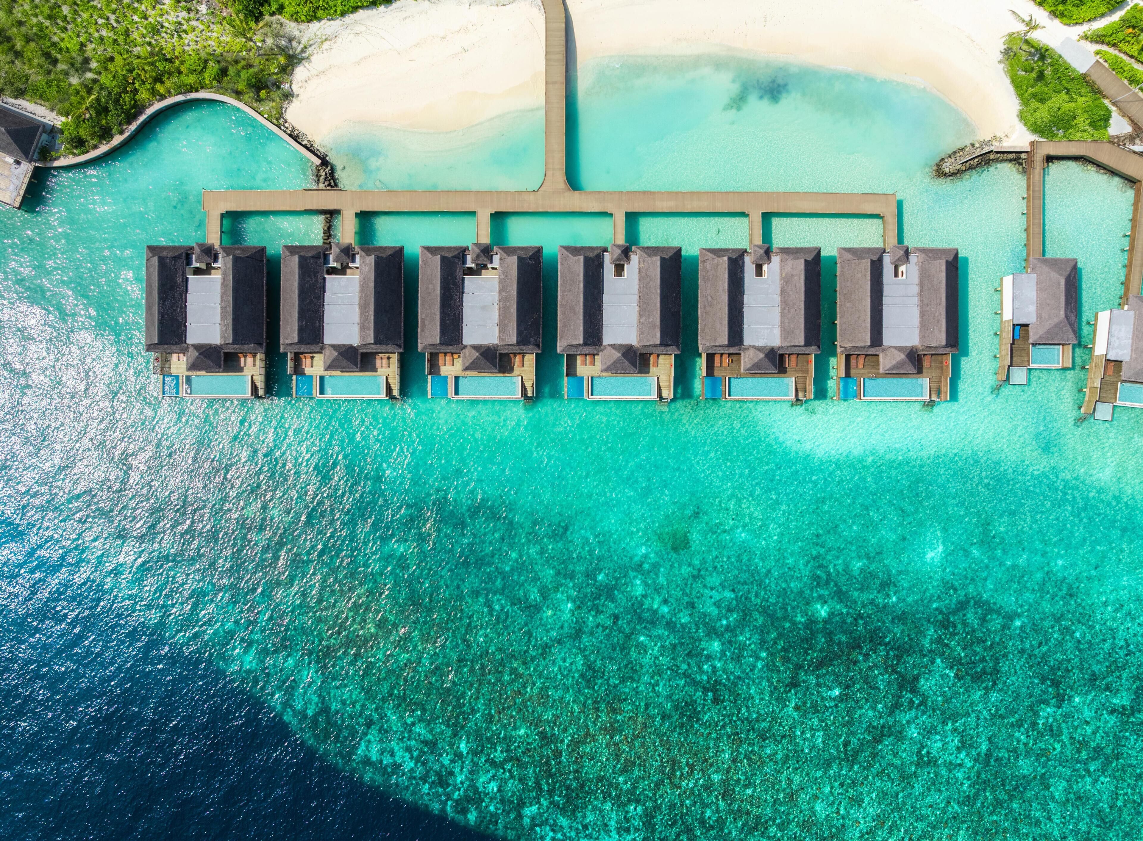 Ett flygbildsbild av ett semesterområde med bungalows som är byggda på pålar över vattnet och som ligger i rad längs en brygga, med anslutning till ett sandstrandområde.
