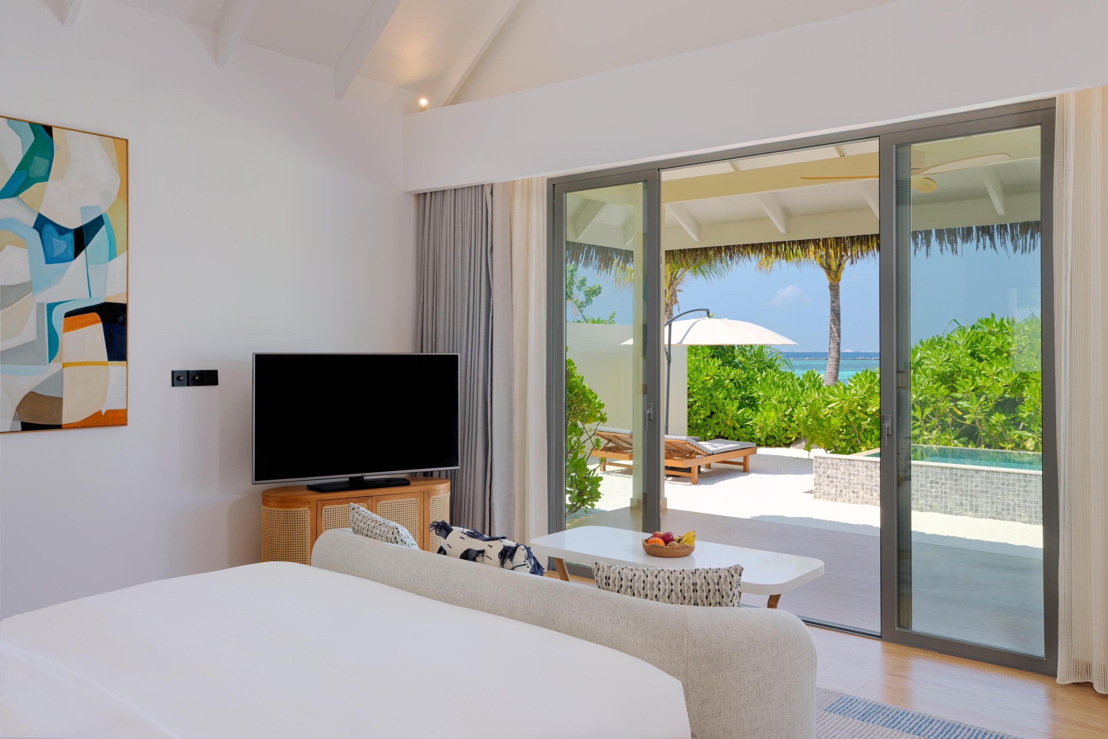 A modern hotel room with a large bed, a flat-screen TV, a dining table with fruit, and a balcony with a pool and tropical view.