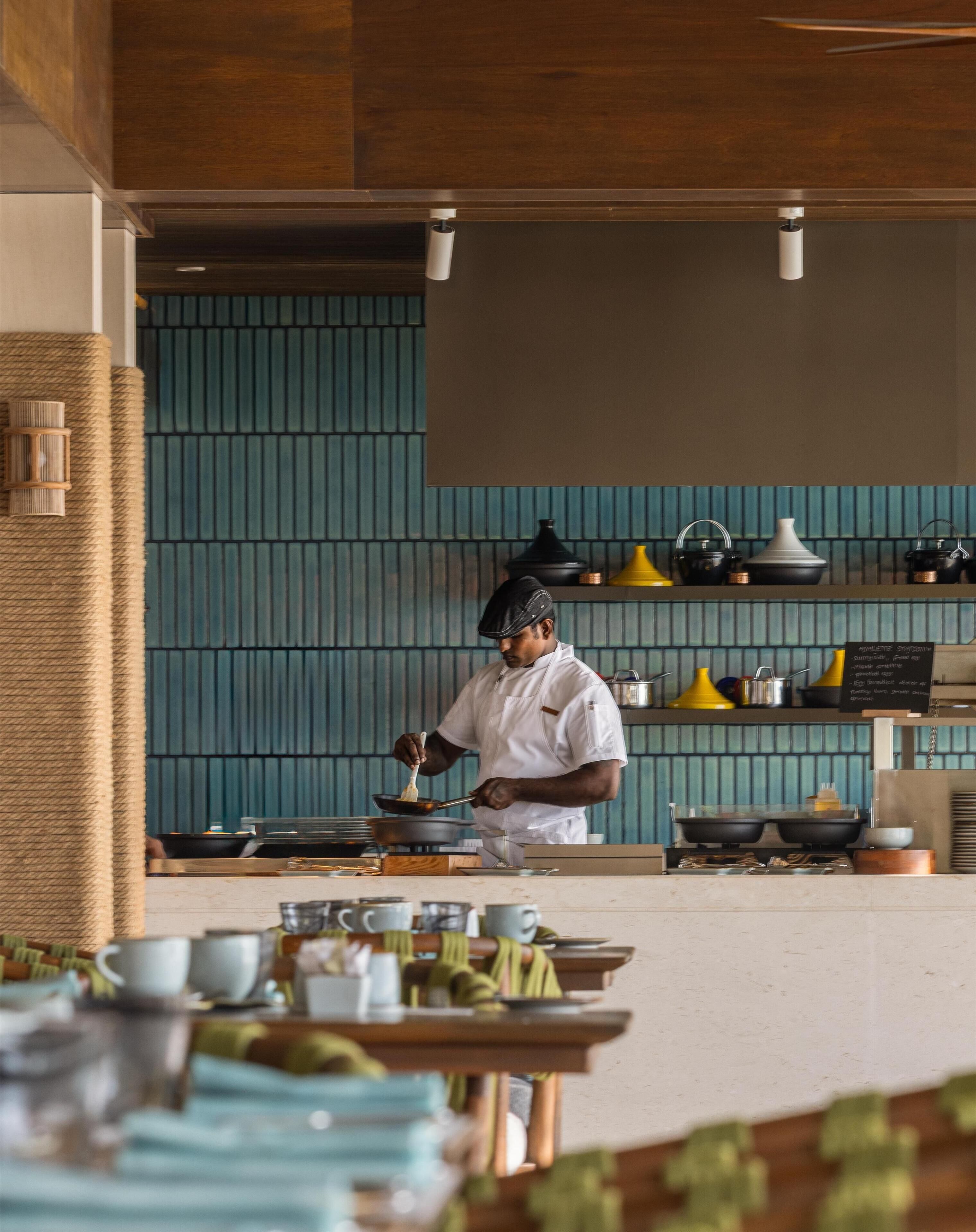 A chef in a white uniform cooking in a modern kitchen with teal walls and various kitchen utensils and appliances.