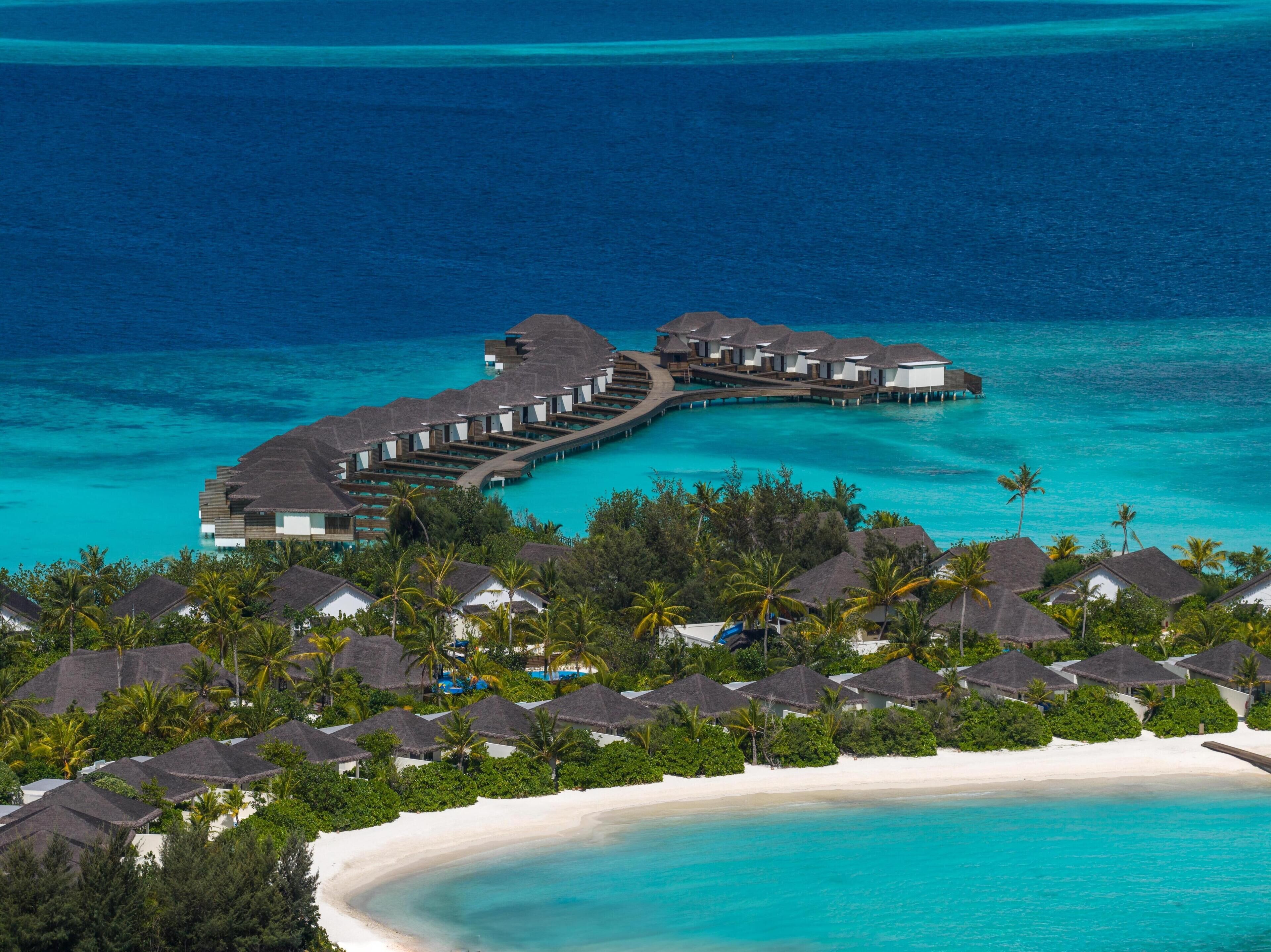 An overwater resort with thatched-roof bungalows connected by walkways, surrounded by lush greenery and a pristine beach.