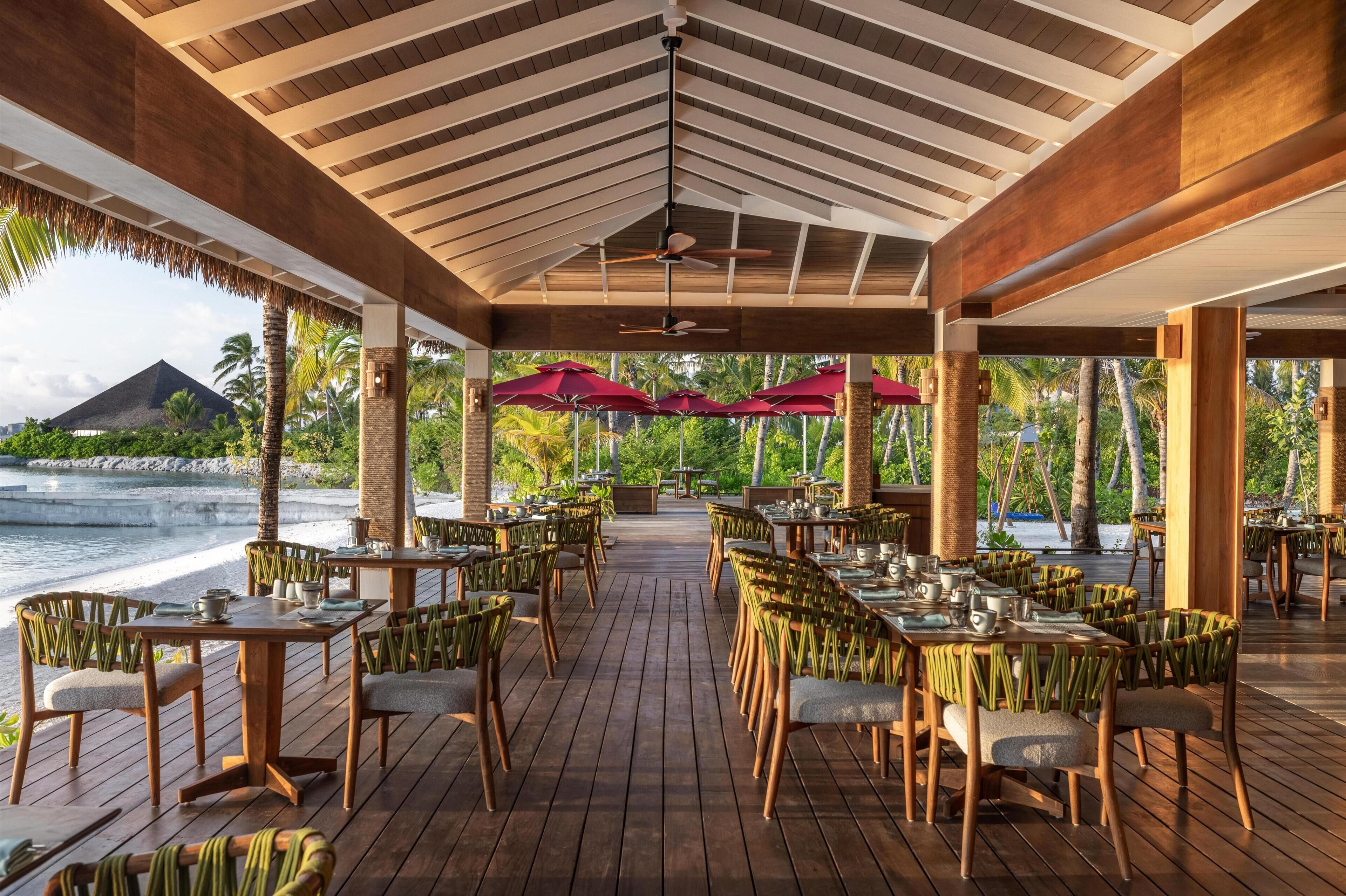 A covered outdoor dining area with wooden tables and chairs, red umbrellas, and a view of the ocean and palm trees.