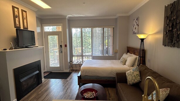 A modern hotel room with a large bed, a sitting area with a sofa, a fireplace, a marble sink, and a view of the outdoors. - Luxe Studio - 102A (Kelowna)