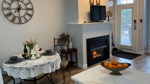 A dining area with a round wooden clock, a fireplace with a TV above it, and a bowl of oranges on a marble countertop. - Luxe Studio - 102A (Kelowna)