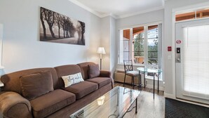 A living room with a brown sofa, a glass coffee table, a painting of trees, a lamp, and a view through a glass door. - Golf Course Walk Up - 102B (Kelowna)