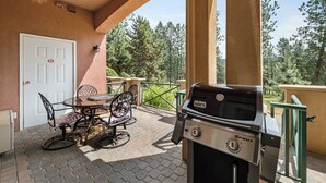 An outdoor patio with a Weber Spirit grill, a dining table with chairs, and a view of trees in the background. - Golf Course Walk Up - 102B (Kelowna)