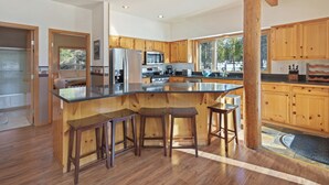 A spacious kitchen with wooden cabinets, a large island with stools, and a view of the outdoors. - 4 Umpqua - Sunny Sunriver! Hot Tub, AC, SHARC, Amazing Location! (Bend)