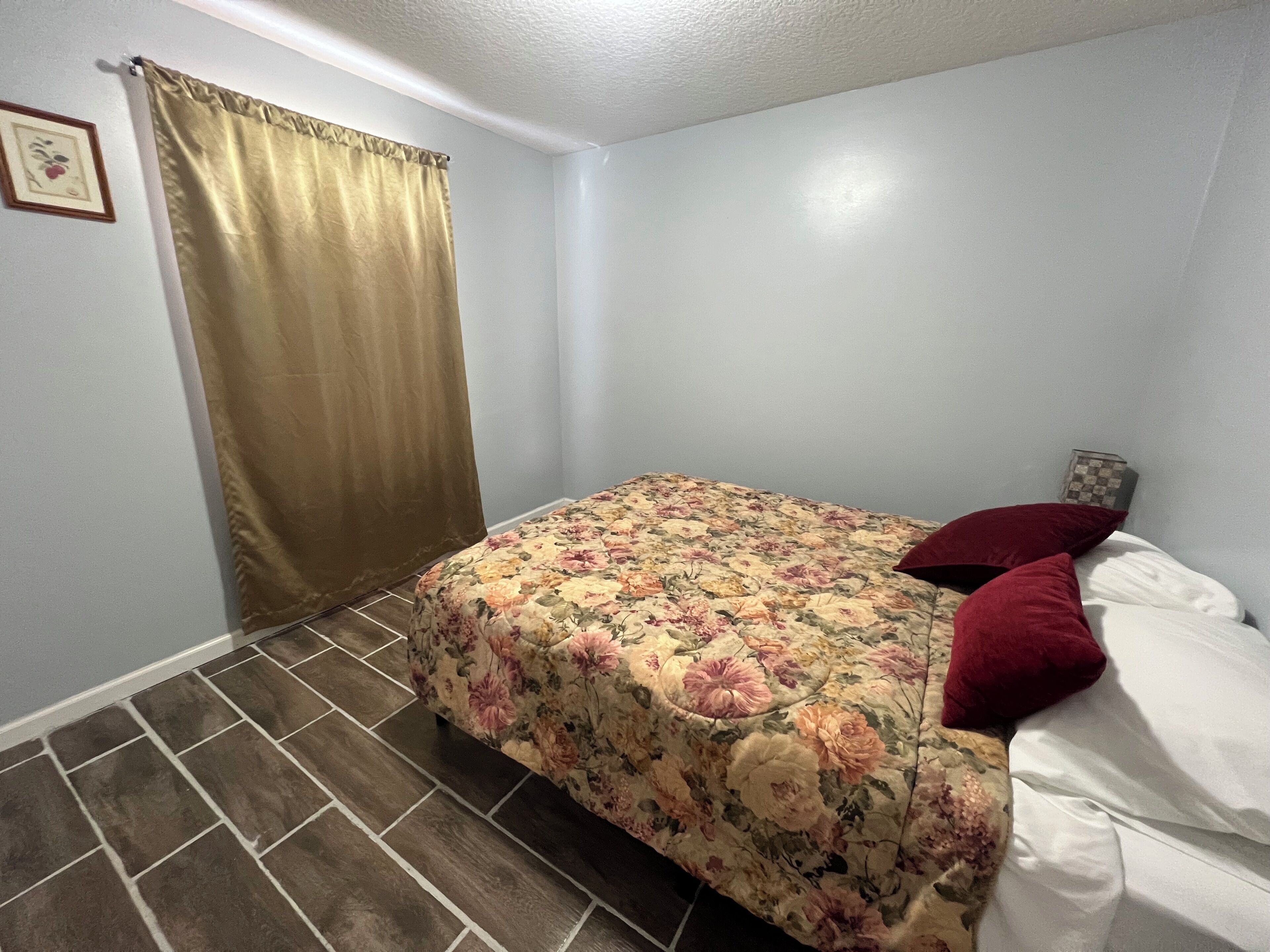 Un dormitorio pequeño con una colcha estampada con motivos florales, una almohada roja y una cortina.