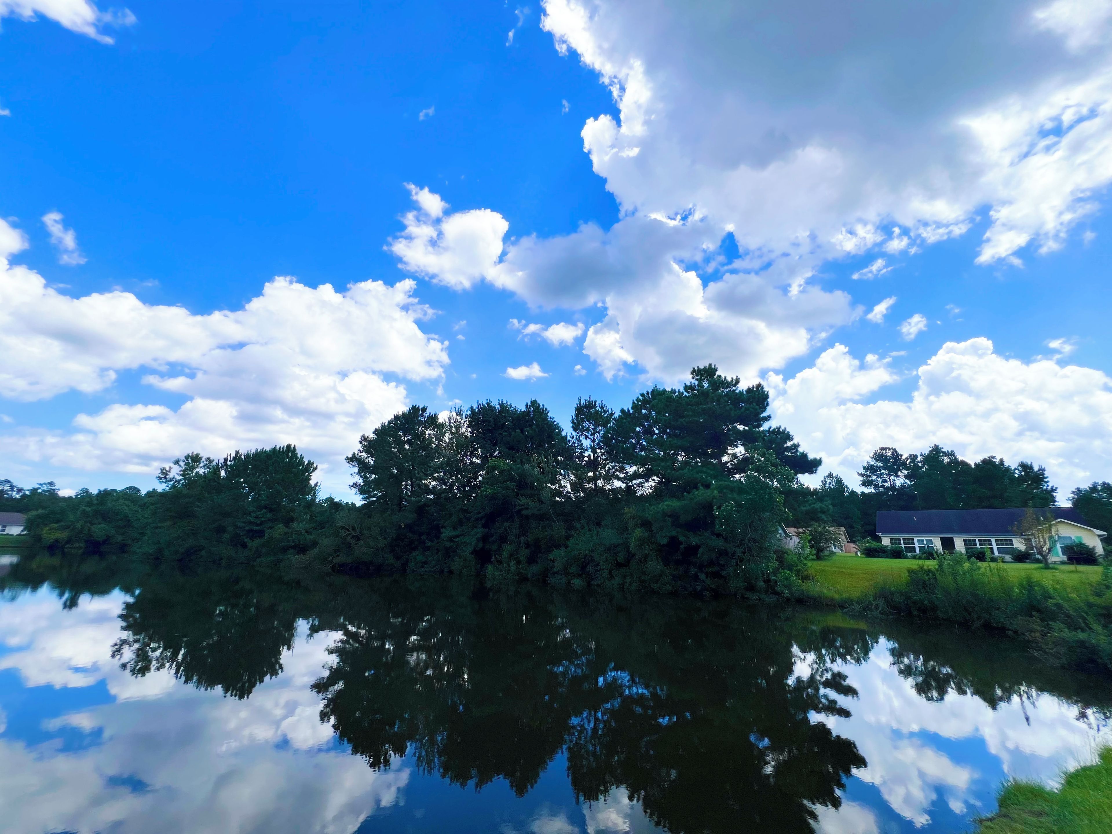Eine friedliche Landschaft mit einem Gewässer, das Himmel und Bäume spiegelt, einem Haus im Hintergrund und freiem Blick auf die umliegende Bepflanzung.