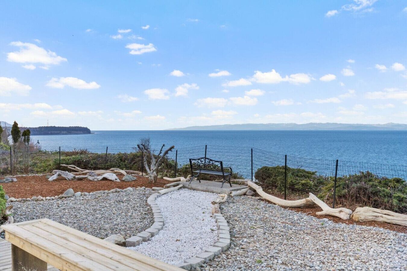 A coastal view with a pebble path, a bench, and a fence overlooking the sea.