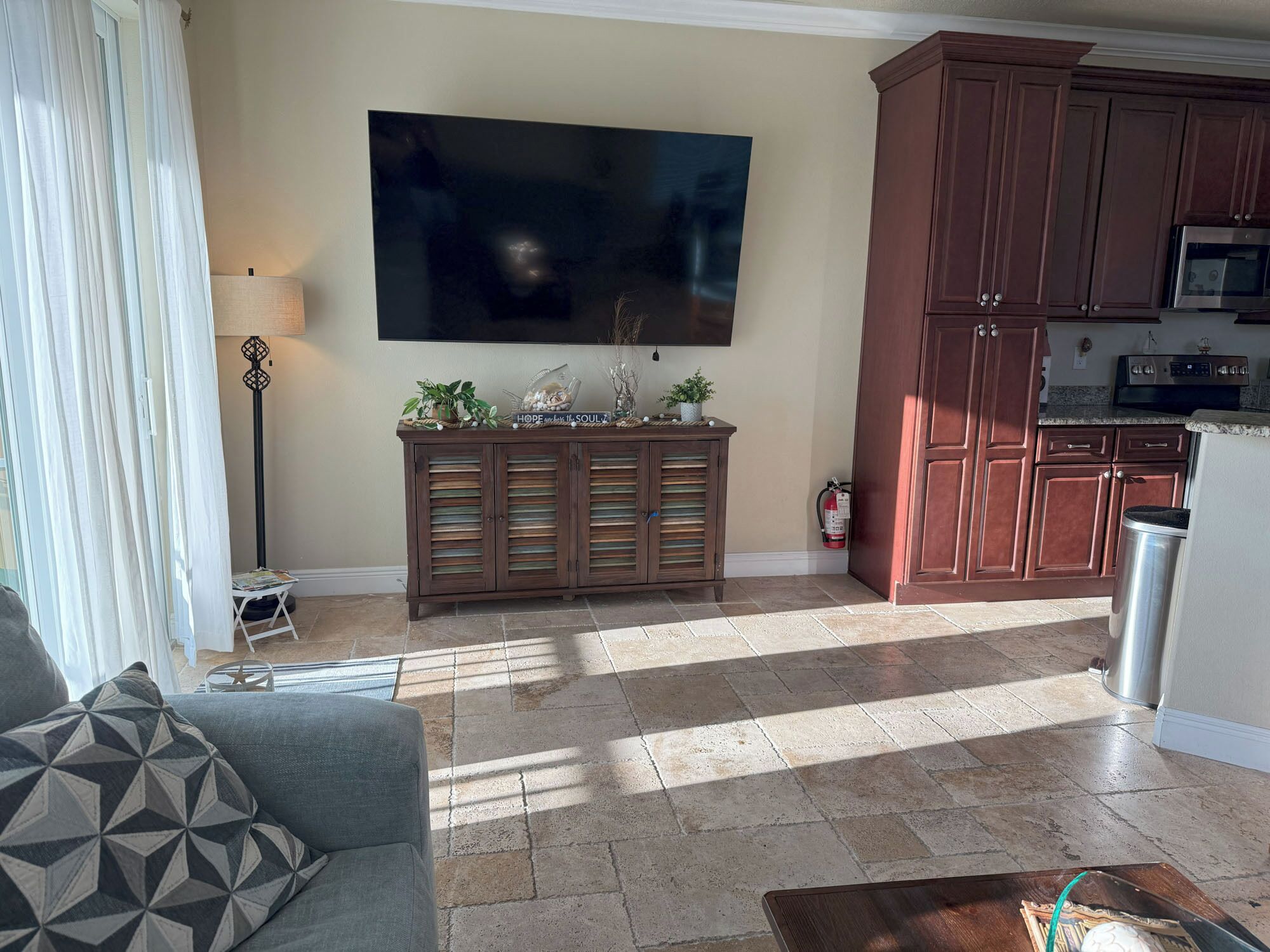 A living room with a sofa, a coffee table, a TV mounted on the wall, and a kitchen area with cabinets and appliances.