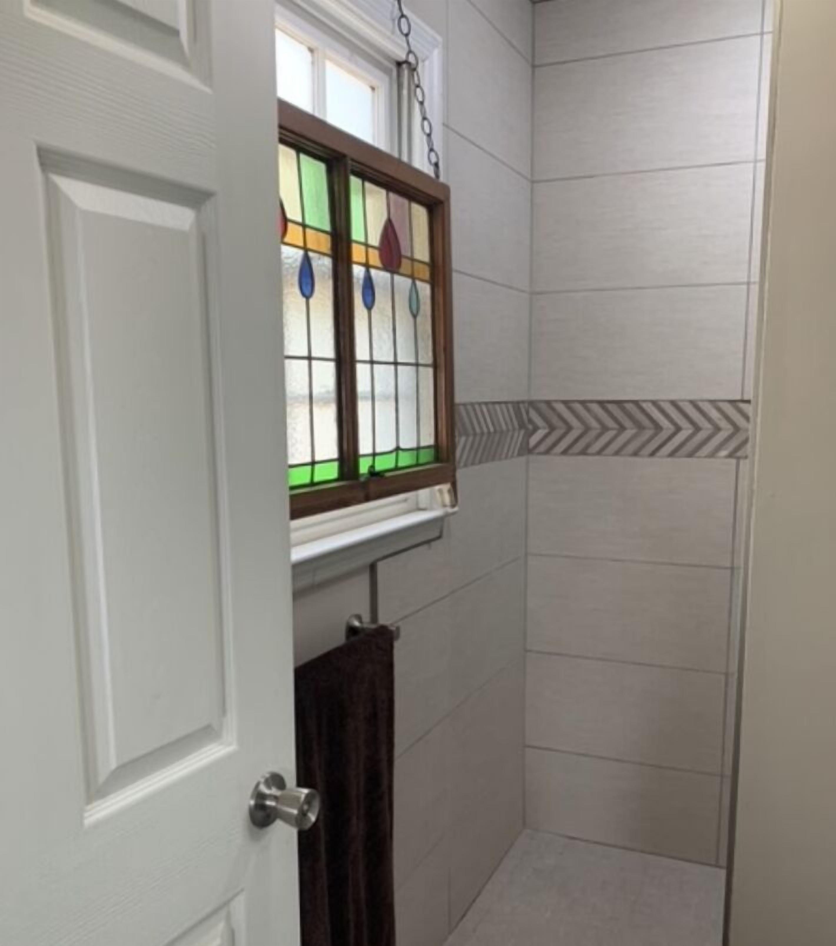 A bathroom with a shower, a door with a towel hanging, and a window with stained glass.
