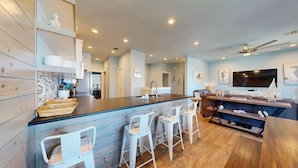 A modern kitchen with a central island, bar stools, a dining area with a TV, and a ceiling fan.