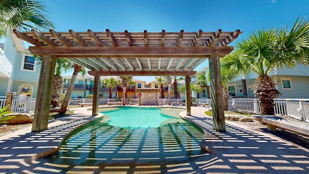 A pool area with a wooden pergola, palm trees, and a building with balconies in the background.