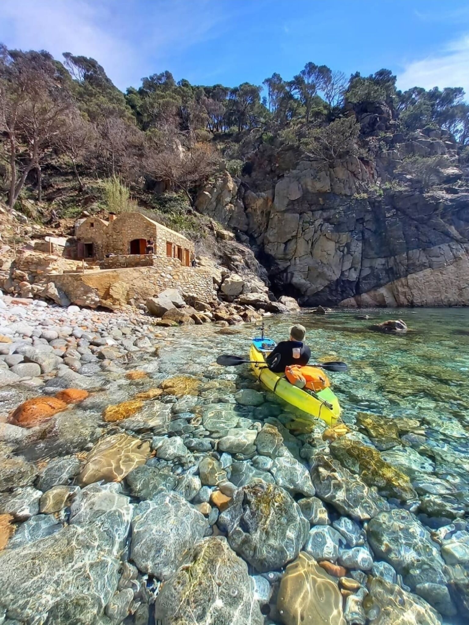 Um praticante de caiaque em uma praia clara e rochosa, com um edifício de pedra próximo.