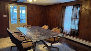A dining area with a glass-top table, four chairs, wooden walls, a ceiling fan, and a window with curtains.