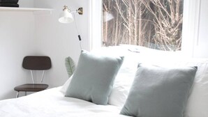 Un dormitorio con cama, dos almohadas, una silla, un escritorio y ventana con vista a árboles.