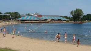 Una playa con gente nadando y caminando por la orilla. Se observa un edificio grande con techo azul al fondo.