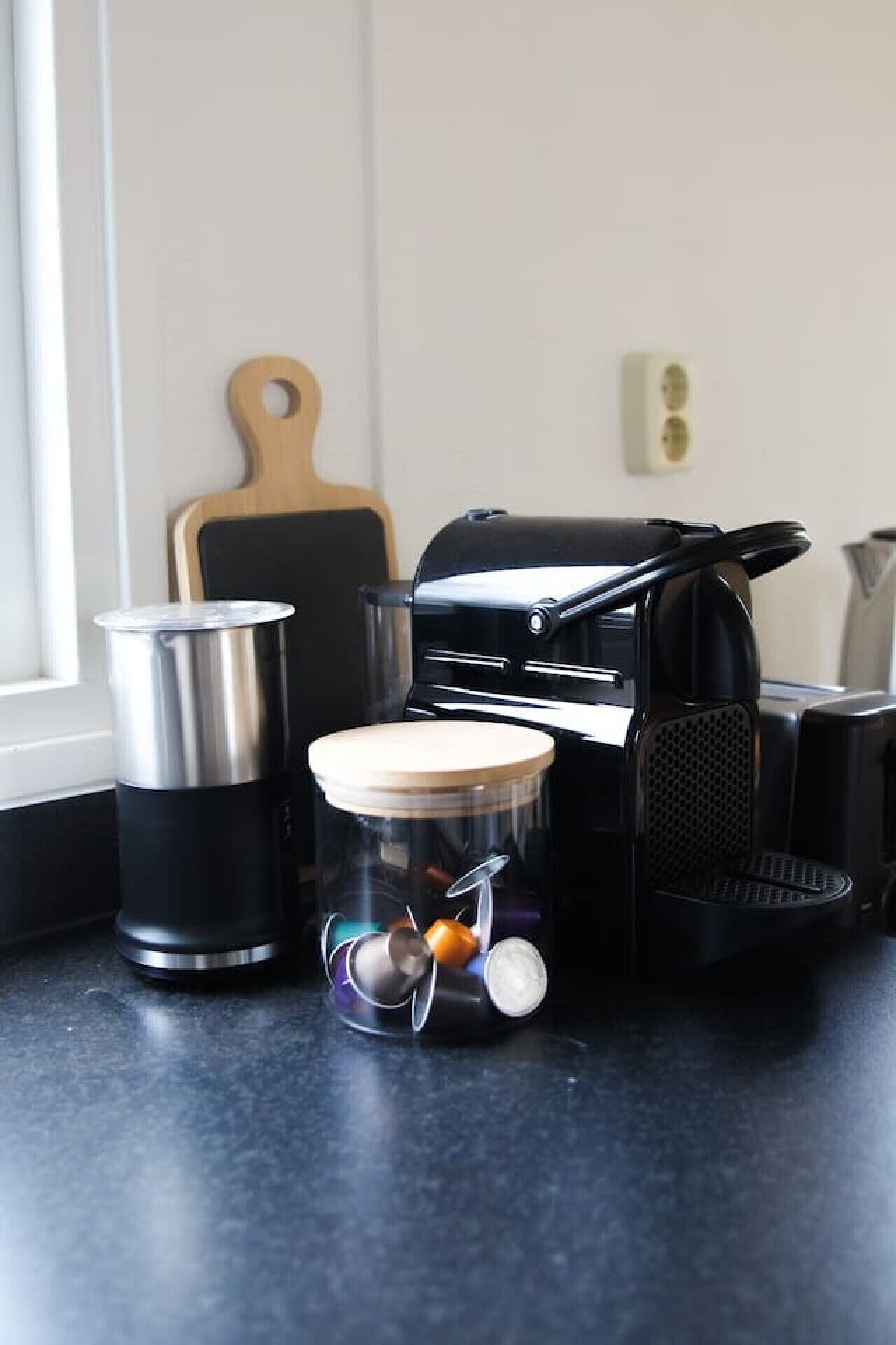 Una encimera de cocina con una cafetera, un recipiente de acero inoxidable, una tabla de cortar de madera y un electrodoméstico negro.