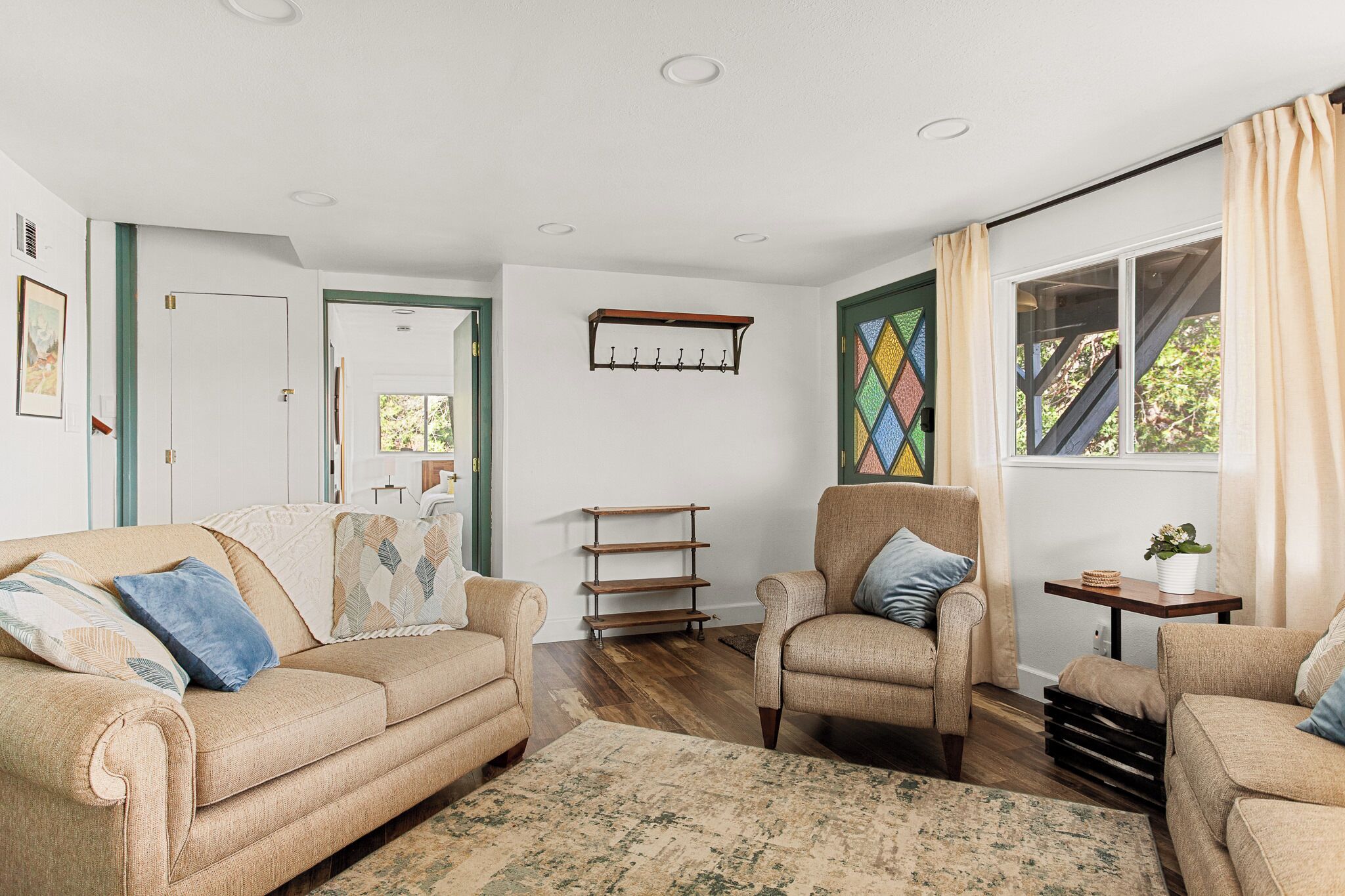 A living room with beige sofas, a patterned rug, wooden shelves, and a window with curtains.