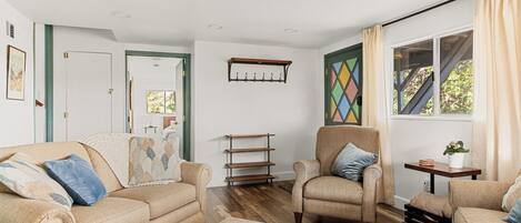 A living room with beige sofas, a patterned rug, wooden shelves, and a window with curtains.