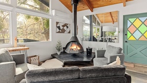 A cozy living room with a stone fireplace, large windows, and a wooden ceiling.