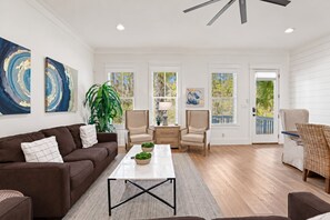 A spacious living room with a brown sofa, a coffee table, and two armchairs. There are large windows with blinds, a potted plant, and artwork on the walls.
