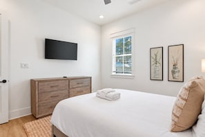 A bedroom with a bed, a wooden dresser, a flat-screen TV, a window with blinds, and two framed pictures on the wall.