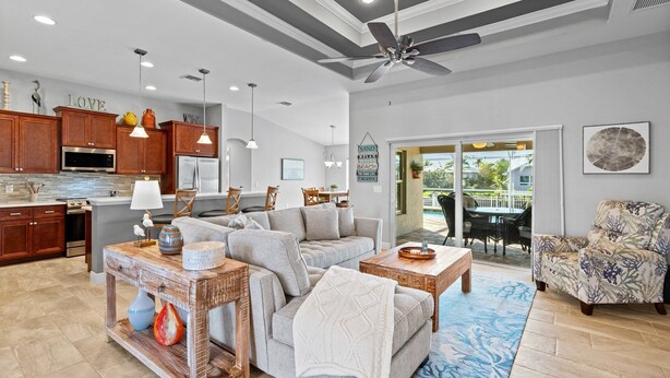 Un'ampia zona giorno con divano, tavolino e oggetti decorativi. C'è una cucina con pensili in legno e un'area pranzo con tavolo e sedie. La stanza dispone di una ventola a soffitto e di vista sull'esterno tramite una porta in vetro.