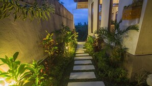 A pathway leading to a building with a concrete wall on one side and a wooden fence on the other, surrounded by lush greenery and plants. - Umah Kemuning Near Central Ubud Bali (Bali)