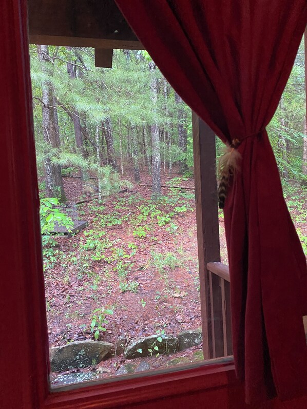 A window with a view of a forested area, curtains partially drawn, and a wooden railing.