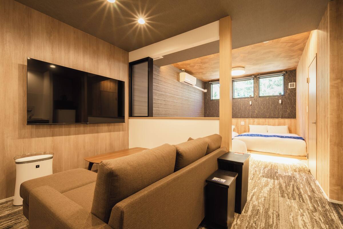 Een moderne hotelkamer met een bank, een klein tafeltje, een aan de muur bevestigde televisie, een bed met een houten hoofdbord en een raam dat natuurlijk licht binnenlaat.