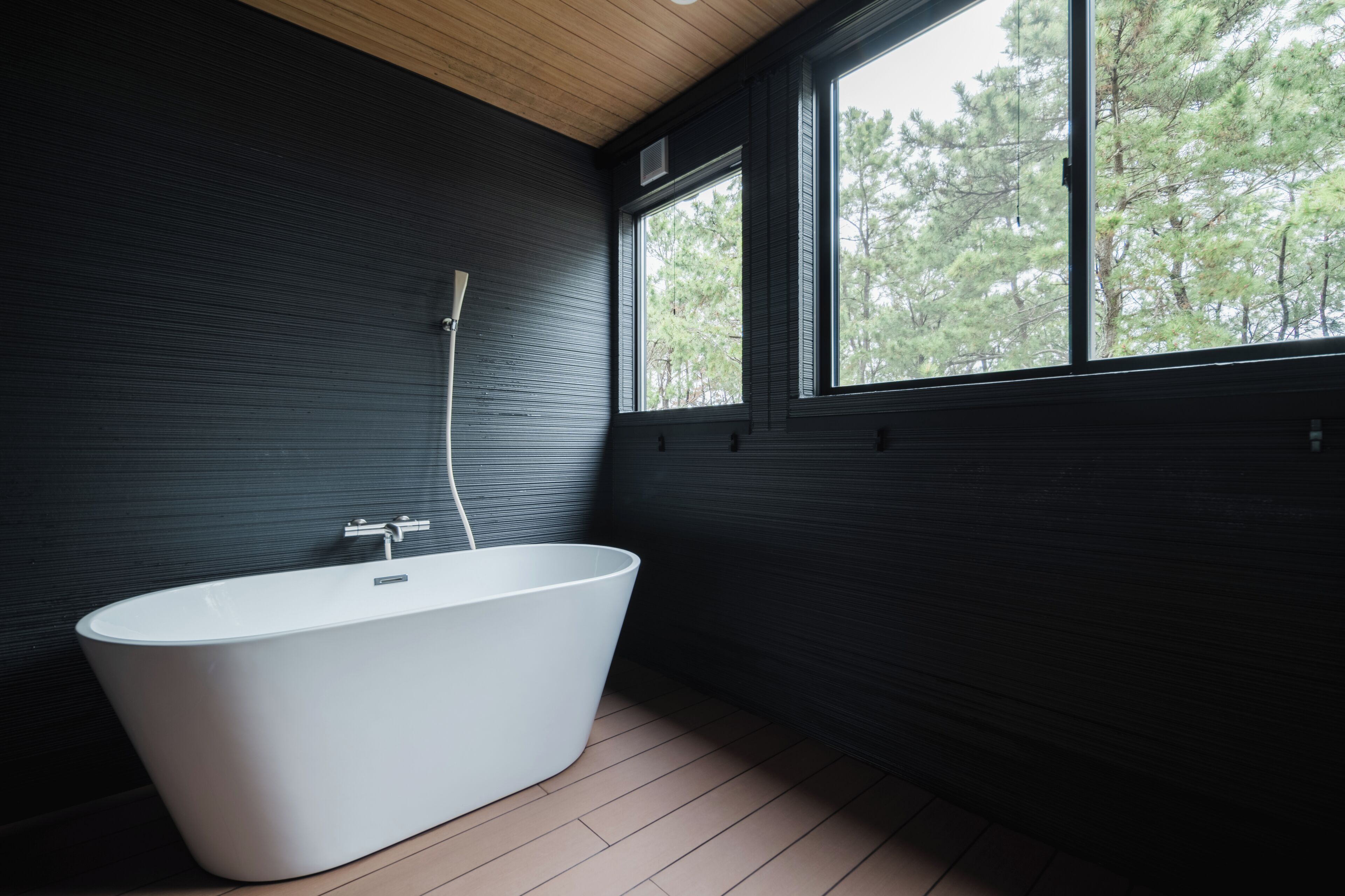 A modern bathroom with a freestanding bathtub, a window with a view of trees, and dark wooden walls.
