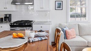 Una cocina moderna con armarios blancos, una campana extractora de acero inoxidable y una estufa negra. Hay una mesa de comedor con un periódico, un cuenco de fruta y una cafetera.