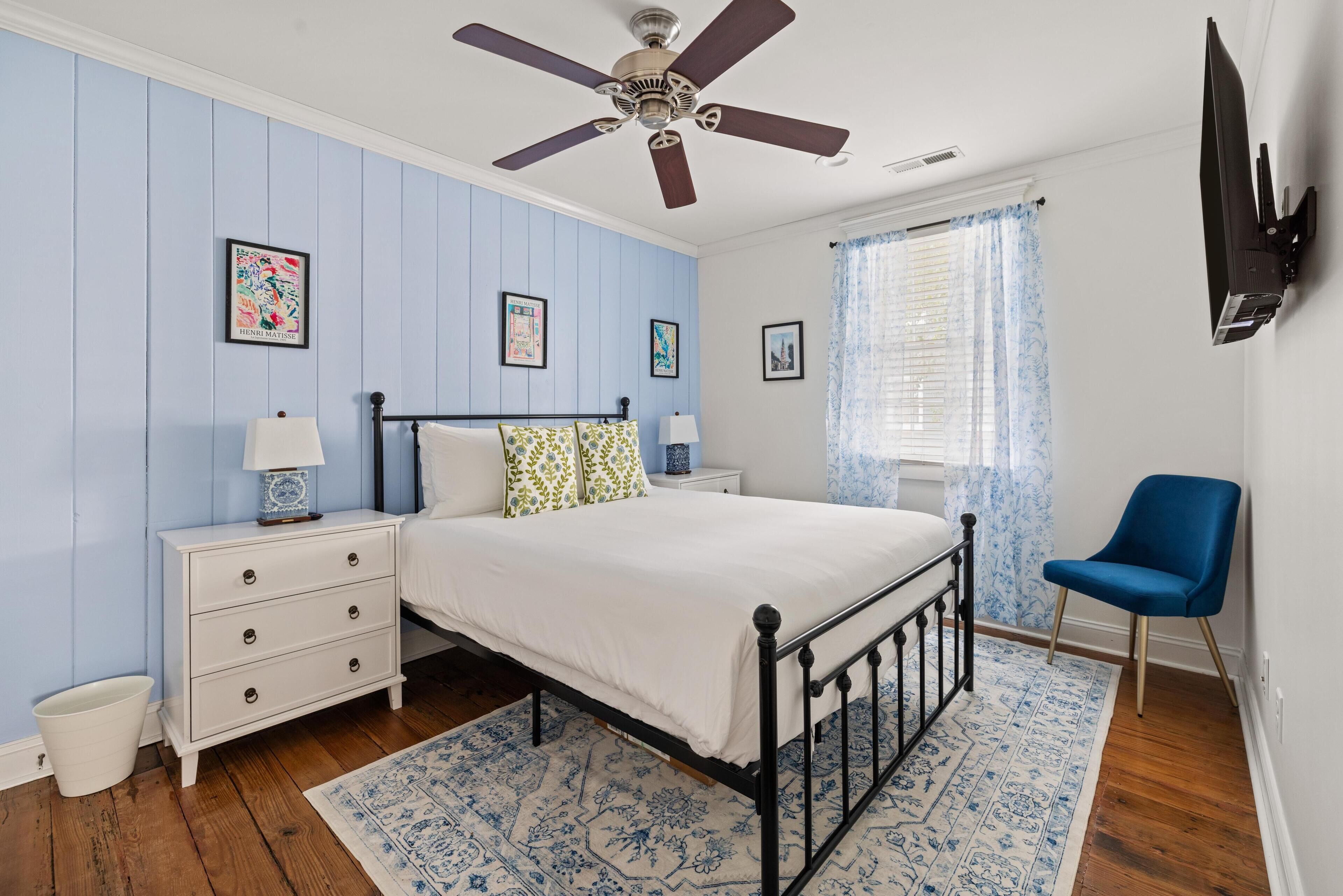 A bedroom with a bed, nightstands, a blue chair, a ceiling fan, and a TV mounted on the wall.