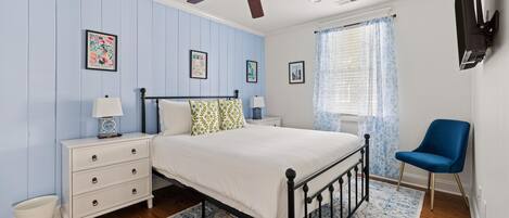 A bedroom with a bed, nightstands, a blue chair, a ceiling fan, and a TV mounted on the wall.