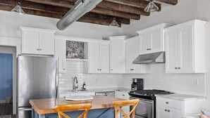 Una cocina con armarios blancos, un refrigerador de acero inoxidable, una isla de madera con dos sillas y una estufa.