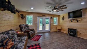 A log cabin interior with a ceiling fan, a mounted television, a fireplace, and a large window.