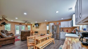 A wooden interior with a dining table, benches, a sofa, a kitchen with appliances, and a mounted deer head.