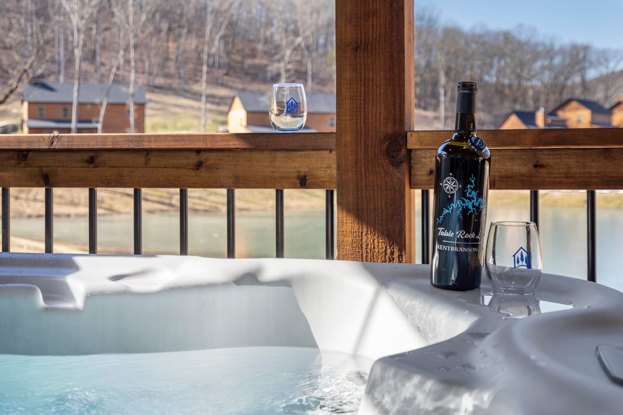 A hot tub with a bottle and glass on a wooden deck overlooking a body of water.