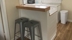 Une petite cuisine avec des meubles blancs, un plan de travail en bois, un évier et deux tabourets.