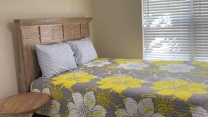 Une chambre avec une tête de lit en bois, une couvre-lit à motifs floraux gris et jaunes, une petite table ronde en bois et un ventilateur de plafond.