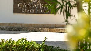 Placa para 'Canaba Beach Club', com cerca de madeira e vegetação ao primeiro plano.
