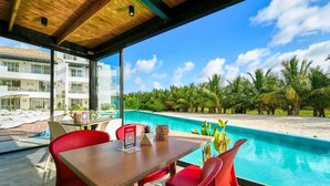 Uma área de refeições com mesa e cadeiras, com vista para uma piscina, palmeiras e um edifício ao fundo.
