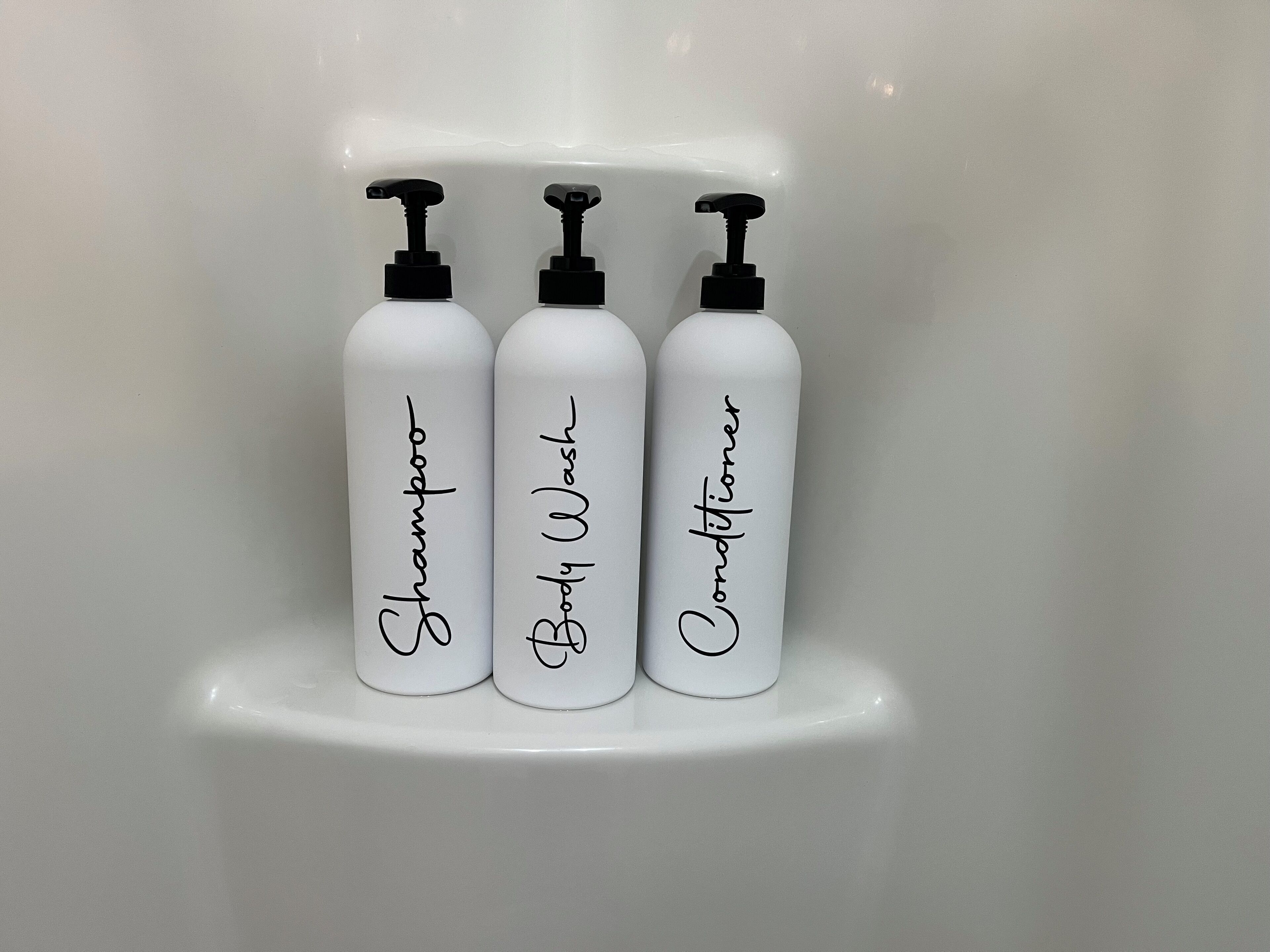 Trois flacons blancs avec des pompes noires, étiquetés respectivement « Shampoing », « Gel douche » et « Après-shampoing », sont posés sur une surface blanche.