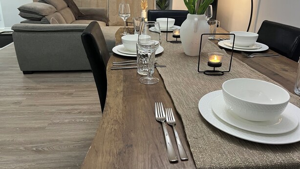 Un comedor con una mesa de madera dispuesta para cuatro personas, con platos, vasos y cubiertos. Hay dos jarrones con flores, una vela y un sofá al fondo.
