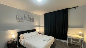 Un dormitorio ordenado con cama, dos mesitas de noche con lámparas, un escritorio con silla y un amplio ventanal con cortinas.