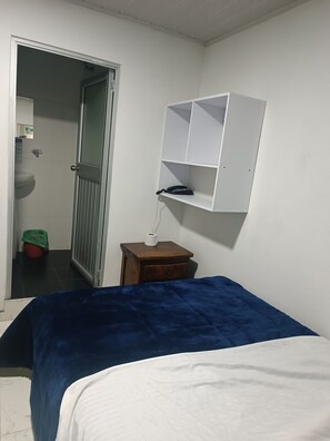A bedroom with a bed, a small wooden nightstand, a white shelf with a telephone and a tissue box, and a door leading to a bathroom. - Hotel The Swit Real Centro (Tunja)