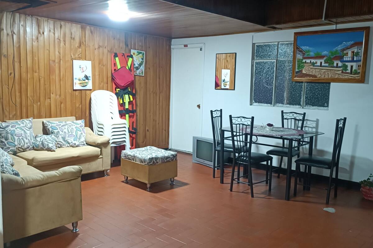 A cozy living room with a wooden ceiling, a sofa, a dining table with chairs, and a TV.