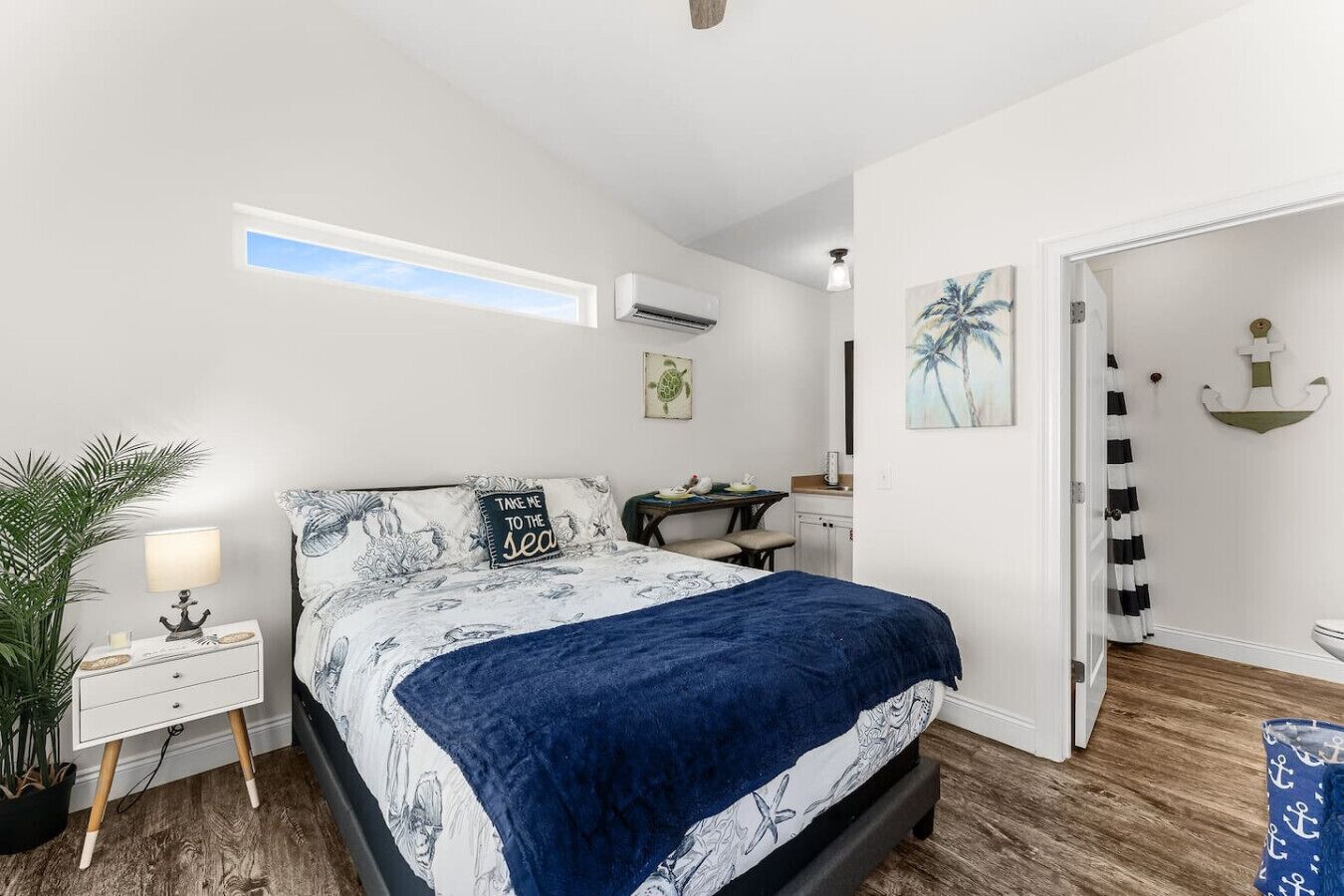 A bedroom with a bed, a desk, a chair, a nightstand, a potted plant, and a door leading to another room.